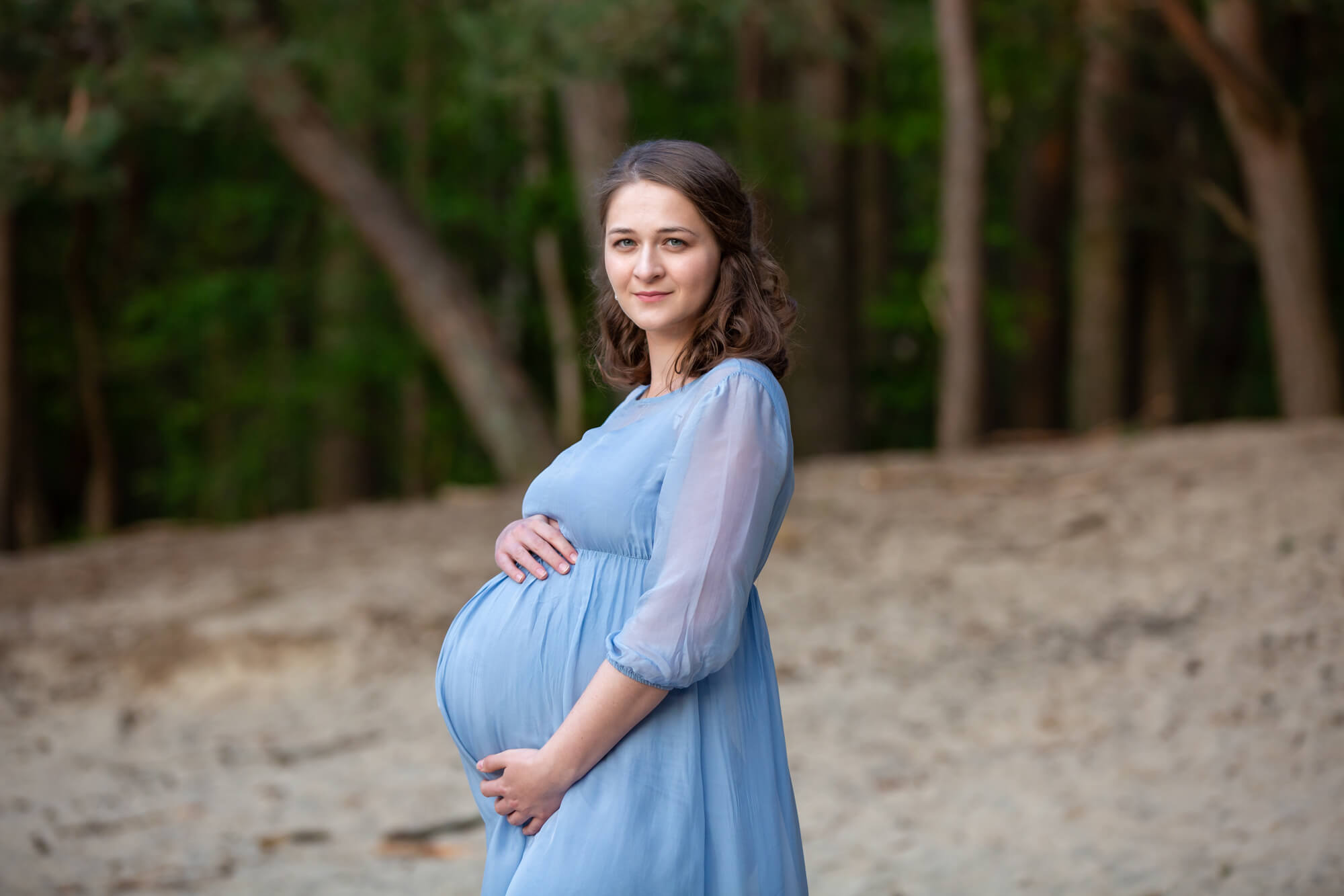 Eine schwangere Frau steht in einem wunderbaren hellblauen Kleid in einem Waldstück bei einem Fotoshooting und streichelt ihren Babybauch.