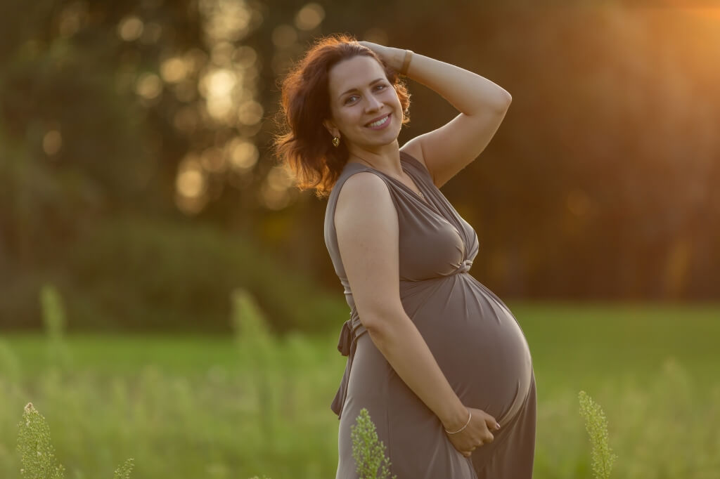 Eine wunderschöne werdende Mama steht beim Sonnenuntergang auf einer grünen Wiese und lächelt posierend.