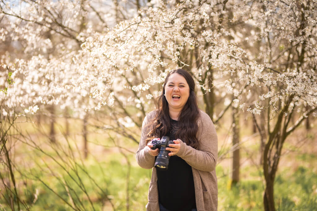 Die Familienfotografin Karola Schnier schaut bei einem Fotoshooting von ihr selbst lachend in die Kamera. Sie steht vor einem blühenden Baum in der Natur.