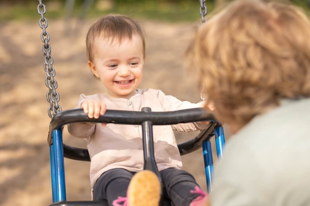 Bei einem Kindershooting auf einem Spielplatz schaukelt eine lachende Zweijährige, während ihre Mama sie dabei fröhlich und lachend anguckt.