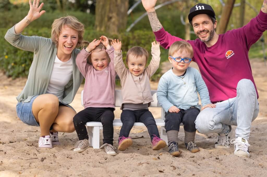 Familienfotografie wird auf diesem Bild sehr deutlich: eine liebende und lachende Familie mit 3 kleinen Kindern sitzt auf einer weißen Kinderbank auf einem Spielplatz und jubelt lachend in die Kamera