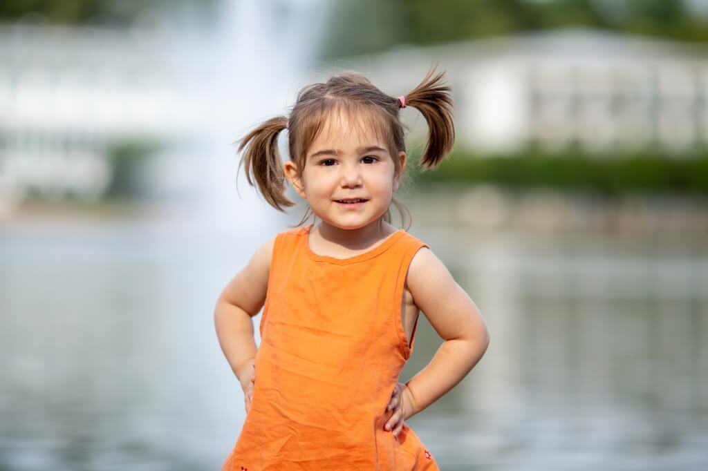 Ein freches Mädchen in einem orangenen Kleid posiert lächelnd vor einem See in Bremen im Rahmen eines Familienshootings.